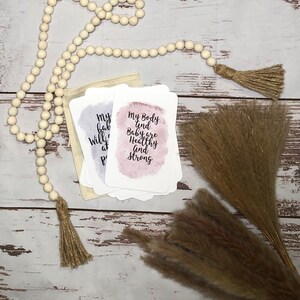 May include: A stack of affirmation cards with watercolor backgrounds and text, including "My body and baby are healthy and strong." The cards are in a small beige pouch, with a wooden bead garland and dried pampas grass.