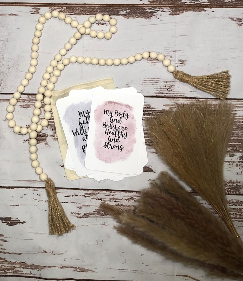 May include: A stack of affirmation cards with watercolor backgrounds and text, including "My body and baby are healthy and strong." The cards are in a small beige pouch, with a wooden bead garland and dried pampas grass.