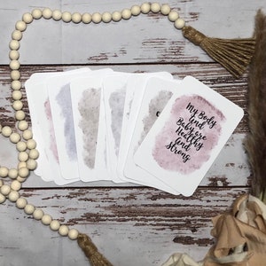 May include: A stack of white affirmation cards with watercolor-style backgrounds in shades of pink, gray, and blue. One card reads "My Body and Baby are Healthy and Strong" in black script. The cards are arranged on a wooden surface with decorative beads and tassels.