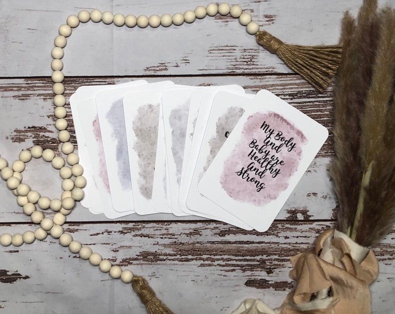 May include: A stack of white affirmation cards with watercolor-style backgrounds in shades of pink, gray, and blue. One card reads "My Body and Baby are Healthy and Strong" in black script. The cards are arranged on a wooden surface with decorative beads and tassels.