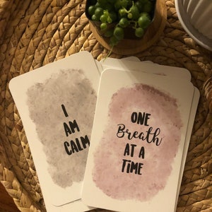 May include: A stack of white affirmation cards with watercolor backgrounds. One card reads "I AM CALM" in black text, and another reads "ONE Breath AT A TIME". The cards are on a woven tray with a small potted plant.