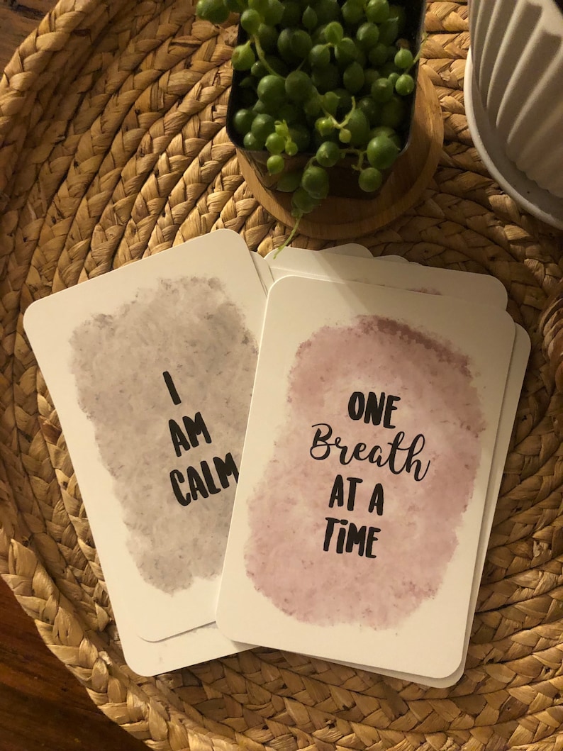 May include: A stack of white affirmation cards with watercolor backgrounds. One card reads "I AM CALM" in black text, and another reads "ONE Breath AT A TIME". The cards are on a woven tray with a small potted plant.