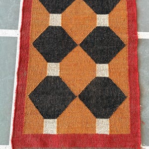 May include: A brown and black geometric patterned rug with a red border and brown fringe. The rug has a diamond pattern with black diamonds on a brown background.