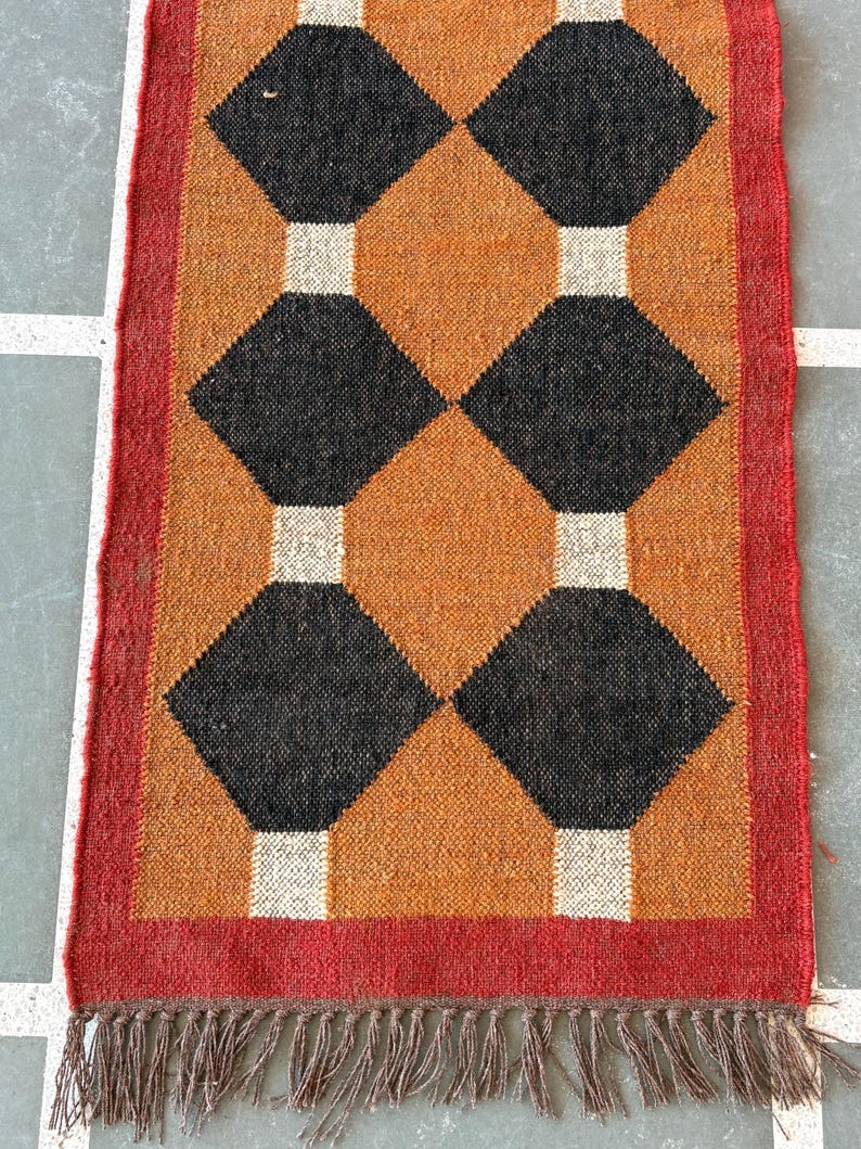 May include: A brown and black geometric patterned rug with a red border and brown fringe. The rug has a diamond pattern with black diamonds on a brown background.