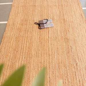 K&ouml;nnte beinhalten: Ein Naturfaser-Teppich aus Jute, einem nachhaltigen und strapazierf&auml;higen Material. Der Teppich hat eine hellbraune Farbe und eine gewebte Textur.