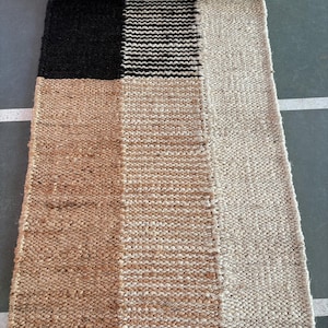 May include: A rectangular jute rug with a geometric pattern. The rug is made of natural fibers and has a woven texture. It features three sections of different colors: black, white, and brown.