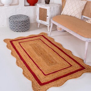 May include: A natural fiber rug with a wavy edge and a red border. The rug is made of jute and has a natural, earthy look.
