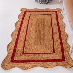 May include: A rectangular jute rug with a scalloped edge and a double red border. The rug is made of natural fibers and has a rustic, earthy look.
