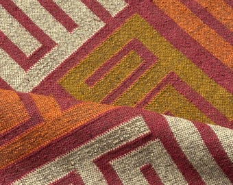 May include: Close-up of a woven textile with a geometric pattern. The design features alternating stripes and angular shapes in shades of red, orange, gold, and off-white. The fabric has a textured appearance, suggesting a natural fiber.