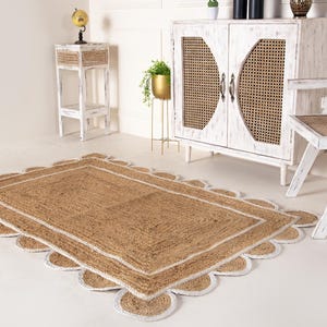 Op de afbeelding: Een vloerkleed van natuurlijke vezels met een witte rand en gegolfde randen. Het vloerkleed ligt op een witte vloer in een kamer met witte meubels.