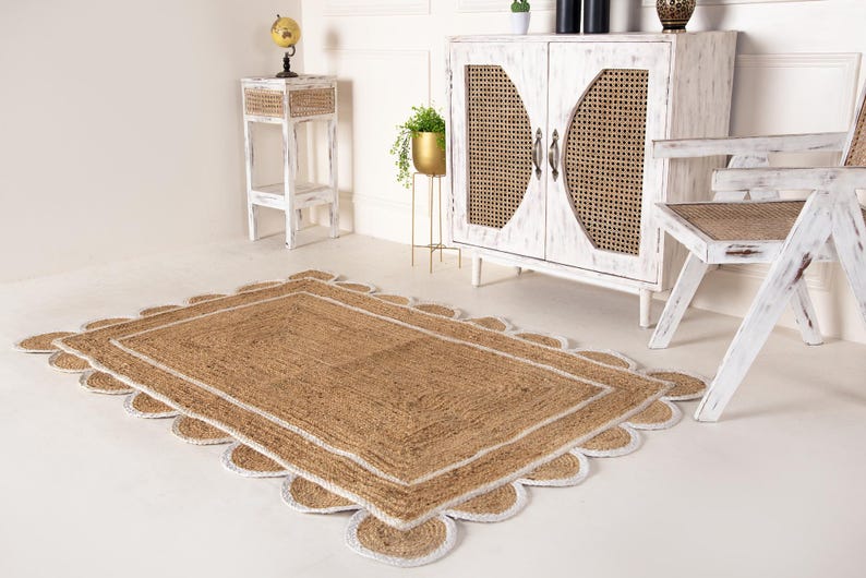 Op de afbeelding: Een vloerkleed van natuurlijke vezels met een witte rand en gegolfde randen. Het vloerkleed ligt op een witte vloer in een kamer met witte meubels.