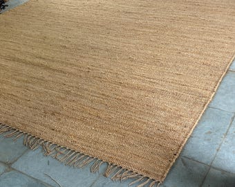 Peut inclure: Un tapis rectangulaire en jute de couleur beige naturel. Le tapis présente une bordure effilochée sur un côté. La texture tissée est visible, avec une surface légèrement irrégulière. Ce tapis convient à un salon ou une chambre, mesurant environ 200 cm x 300 cm.
