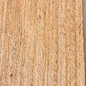 K&ouml;nnte beinhalten: Ein Naturfaser-Teppich aus Jute, einem gewebten Material in hellbrauner Farbe. Der Teppich hat eine geflochtene Textur und ein schlichtes Design.