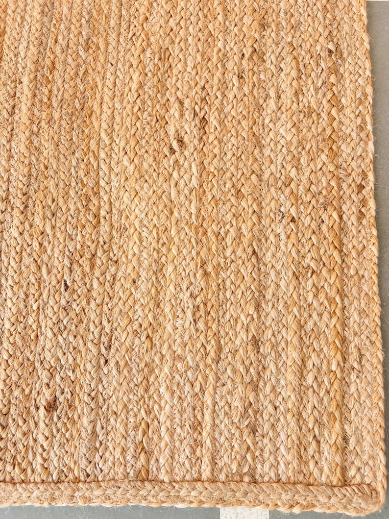 K&ouml;nnte beinhalten: Ein Naturfaser-Teppich aus Jute, einem gewebten Material in hellbrauner Farbe. Der Teppich hat eine geflochtene Textur und ein schlichtes Design.