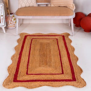 May include: A natural fiber rug with a red border and a wavy edge. The rug is made of jute and has a natural, earthy color.