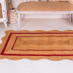 May include: A rectangular jute rug with a red border and a wavy edge. The rug has a natural, earthy tone and is perfect for adding a touch of warmth and texture to any room.