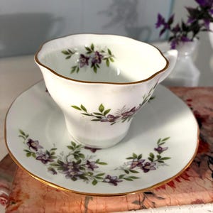 May include: White teacup and saucer set with a floral pattern of purple flowers and green leaves. The rim of the cup and saucer are trimmed with gold. The teacup has a scalloped edge.