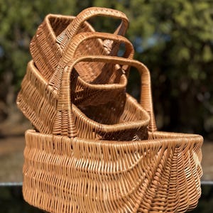 May include: A stack of three woven wicker baskets with arched handles. The baskets are a warm brown color and have a textured surface. The largest basket is at the bottom, with two smaller ones stacked on top.