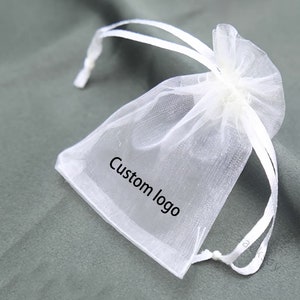 Puede incluir: Una bolsa blanca transparente de organza con cordón y el texto "Custom logo" impreso en la parte delantera. La bolsa está atada con una cinta de raso blanca. La bolsa se utiliza probablemente para empaquetar pequeños regalos o joyas.