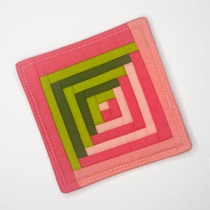 May include: A square fabric coaster with a pink and green log cabin pattern. The coaster is made of multiple layers of fabric sewn together to create a quilted effect.