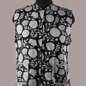 Op de afbeelding: Een zwart-wit vest met bloemenprint, een opstaande kraag en twee voorzakken. Het mouwloze vest heeft een gewatteerde textuur. Het bloemenpatroon bevat grote, gestileerde bloemen en bladeren.