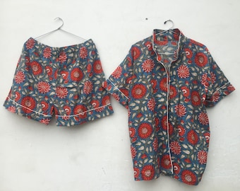 May include: A matching pajama set featuring a short-sleeved button-up shirt and shorts. The set is adorned with a floral pattern in red and white on a blue background. The edges are trimmed with white piping.