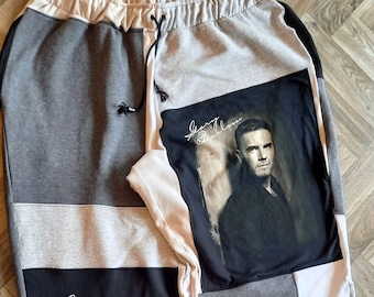 May include: Gray sweatpants with a patchwork design featuring black, white, and gray fabric. The pants have a drawstring waist and a Gary Barlow concert poster on the right leg. The text "in concert in support of young people" is printed on the left leg. The pants have a "Woo's Fashion" logo on the bottom left leg.