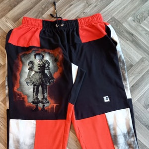 May include: Red, black and white sweatpants with a graphic print of Pennywise the Dancing Clown from the film "IT". The sweatpants have a drawstring waist and a logo on the right leg.