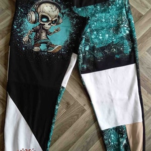 Puede incluir: Pantalones de chándal negros con un estampado gráfico de una calavera de dibujos animados con auriculares y una chaqueta. Los pantalones de chándal tienen una combinación de colores turquesa, blanco y beige.