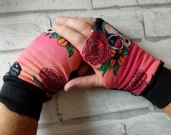 May include: Pink and black fingerless gloves with a floral and skull design. The gloves feature a vibrant pink background with red flowers, green leaves, and a skull with scissors. The cuffs are solid black.