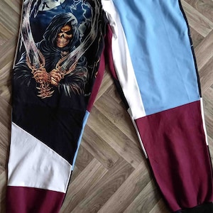 Op de afbeelding: Zwarte sweatpants met een kleurrijk patchwork-ontwerp. Eén been heeft een grafische print van een gekapte figuur die twee zwaarden vasthoudt. De sweatpants hebben een zwart trekkoord en een wit en zwarte tailleband.