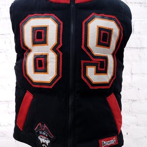 Könnte beinhalten: Schwarze Weste mit der Nummer 89 in Weiß, Orange und Rot. Die Weste hat einen roten Kragen und Akzente an den Taschen. Ein Piratenlogo-Patch und das Wort "Buccaneers" sind sichtbar.
