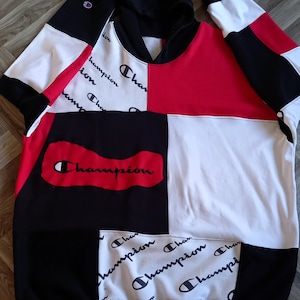Op de afbeelding: Een zwart, wit en rood hooded sweatshirt met een patchwork ontwerp. De trui heeft het woord "Champion" in een rood, wit en zwart kleurenschema.