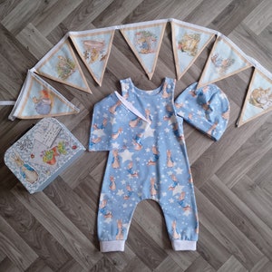 May include: A blue baby onesie with a white star and Peter Rabbit print. A matching blue and white hat and a Peter Rabbit themed banner are also included.