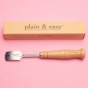 May include: A wooden bread lame with a stainless steel blade and a brown leather cover. The lame is labeled "plain & easy" and is on a pink background.