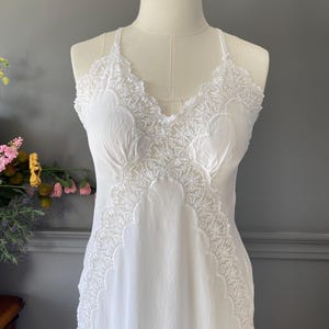May include: A white sleeveless slip dress featuring intricate lace trim along the V-neckline and down the front panels. The garment has thin spaghetti straps and a smooth, lightweight fabric construction that drapes down from the bodice.