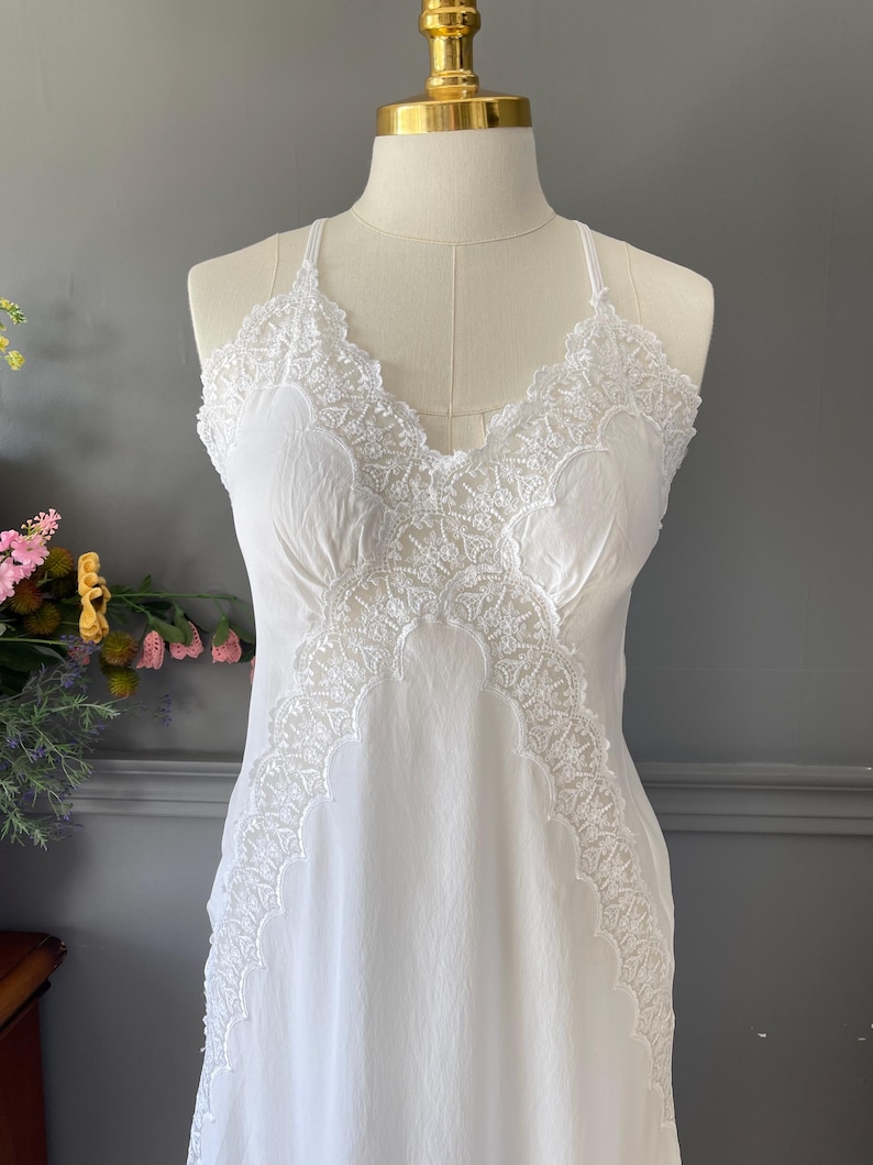 May include: A white sleeveless slip dress featuring intricate lace trim along the V-neckline and down the front panels. The garment has thin spaghetti straps and a smooth, lightweight fabric construction that drapes down from the bodice.