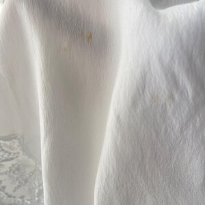 May include: A white fabric with a textured, slightly crinkled surface and a delicate lace trim along the bottom edge. The material features two small, faint, light brown spots visible on the draped folds of the cloth.