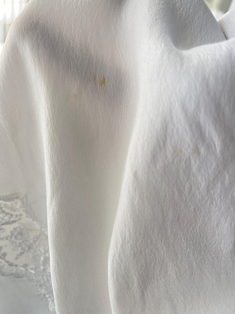 May include: A white fabric with a textured, slightly crinkled surface and a delicate lace trim along the bottom edge. The material features two small, faint, light brown spots visible on the draped folds of the cloth.