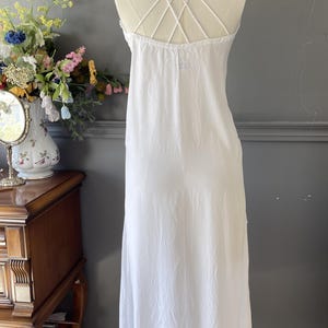May include: A white sleeveless dress featuring a gathered neckline and multiple thin spaghetti straps that cross over the back in a crisscross pattern. The dress has a loose, flowing silhouette that extends to a mid-length hemline.