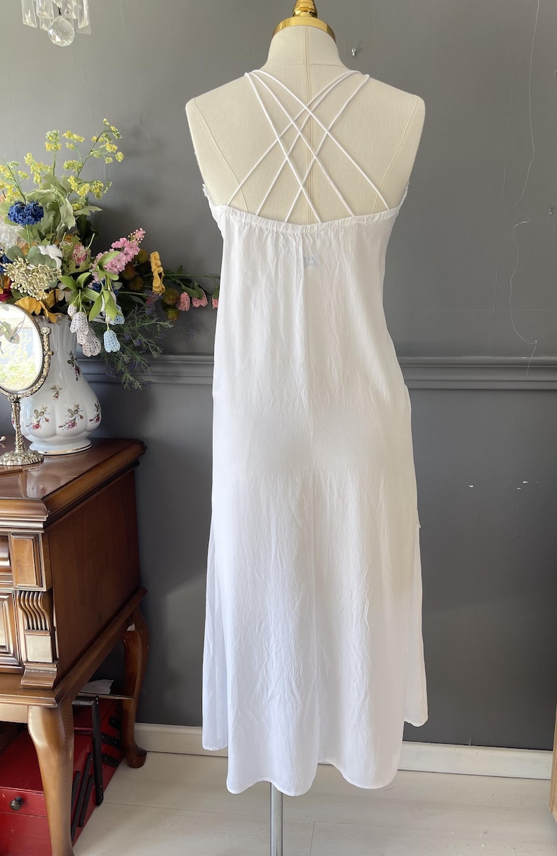 May include: A white sleeveless dress featuring a gathered neckline and multiple thin spaghetti straps that cross over the back in a crisscross pattern. The dress has a loose, flowing silhouette that extends to a mid-length hemline.