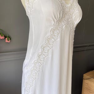 May include: A white sleeveless nightgown featuring delicate lace trim along the neckline and a diagonal lace panel extending down the front. The garment is made of a smooth, lightweight fabric and has thin shoulder straps, showcasing a clean design.
