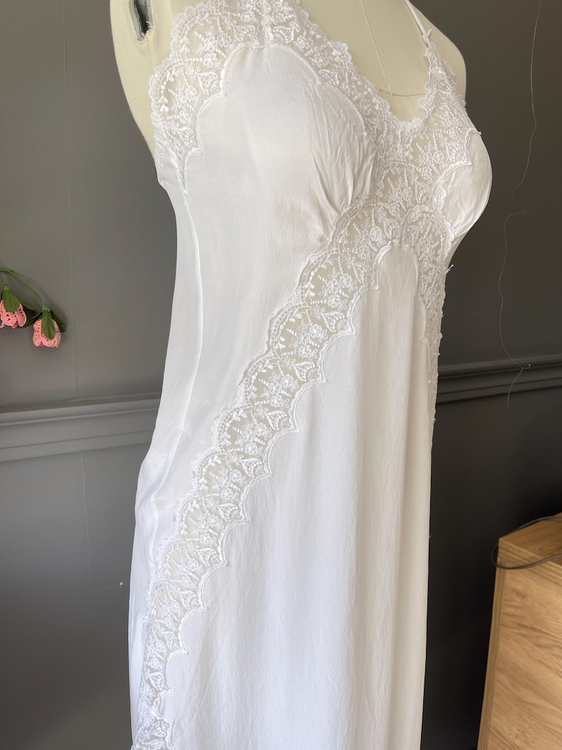 May include: A white sleeveless nightgown featuring delicate lace trim along the neckline and a diagonal lace panel extending down the front. The garment is made of a smooth, lightweight fabric and has thin shoulder straps, showcasing a clean design.