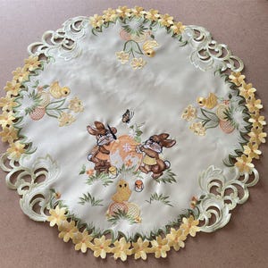 May include: A white embroidered table runner with a scalloped edge features a design of bunnies painting Easter eggs, chicks, and flowers.