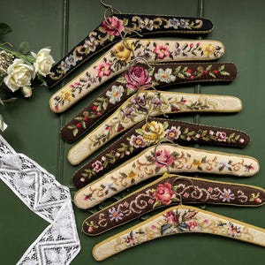 Op de afbeelding: Een verzameling decoratieve kledinghangers met bloemenborduurwerk. De hangers hebben een verscheidenheid aan kleuren, waaronder zwart, bruin en crème, met geborduurde bloemen in roze, geel en blauw. De hangers zijn gerangschikt op een groene ondergrond.