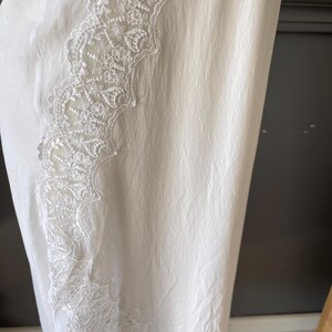 May include: A white garment featuring a textured, slightly crinkled fabric surface. A vertical panel of intricate, floral-patterned white lace trim runs along the side, showcasing detailed embroidery and scalloped edges against the plain fabric.