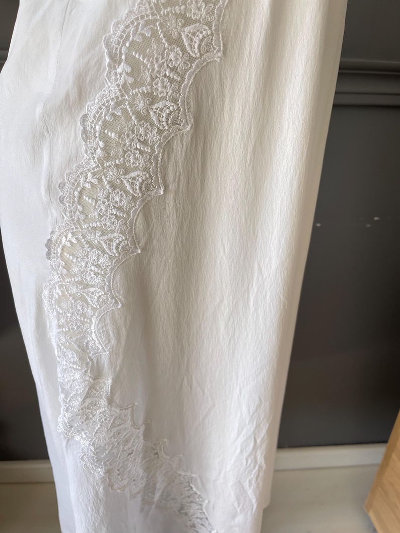 May include: A white garment featuring a textured, slightly crinkled fabric surface. A vertical panel of intricate, floral-patterned white lace trim runs along the side, showcasing detailed embroidery and scalloped edges against the plain fabric.