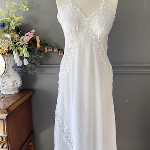 May include: A white sleeveless nightgown featuring a V-neckline with intricate lace trim along the bodice and a diagonal lace panel extending down the side. The garment has thin shoulder straps and a flowing, floor-length skirt silhouette.