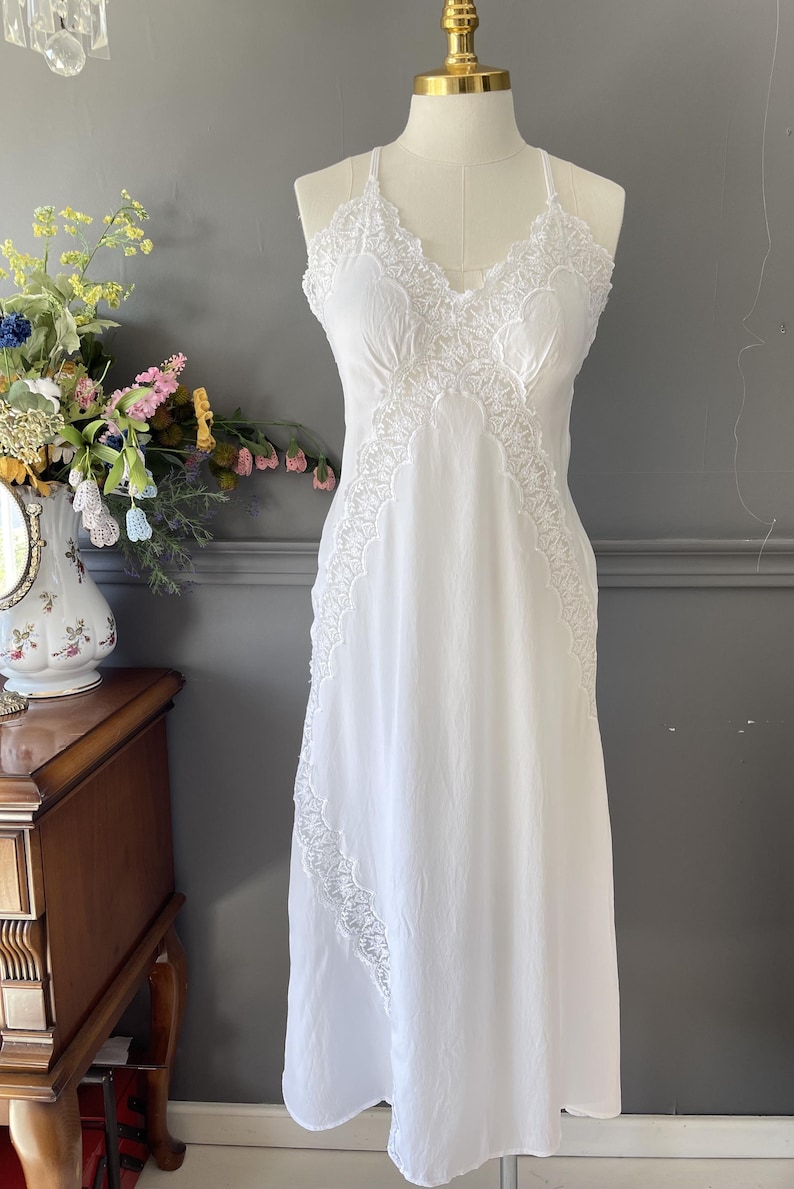 May include: A white sleeveless nightgown featuring a V-neckline with intricate lace trim along the bodice and a diagonal lace panel extending down the side. The garment has thin shoulder straps and a flowing, floor-length skirt silhouette.