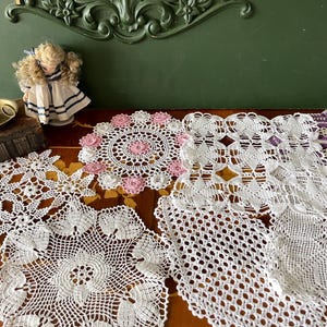 Könnte beinhalten: Fünf weiße Häkeldeckchen mit verschiedenen Mustern, darunter ein rundes Deckchen mit rosa Blüten, ein quadratisches Deckchen mit geometrischem Muster und ein Deckchen mit Blumenmuster. Die Deckchen sind auf einer Holzoberfläche angeordnet.